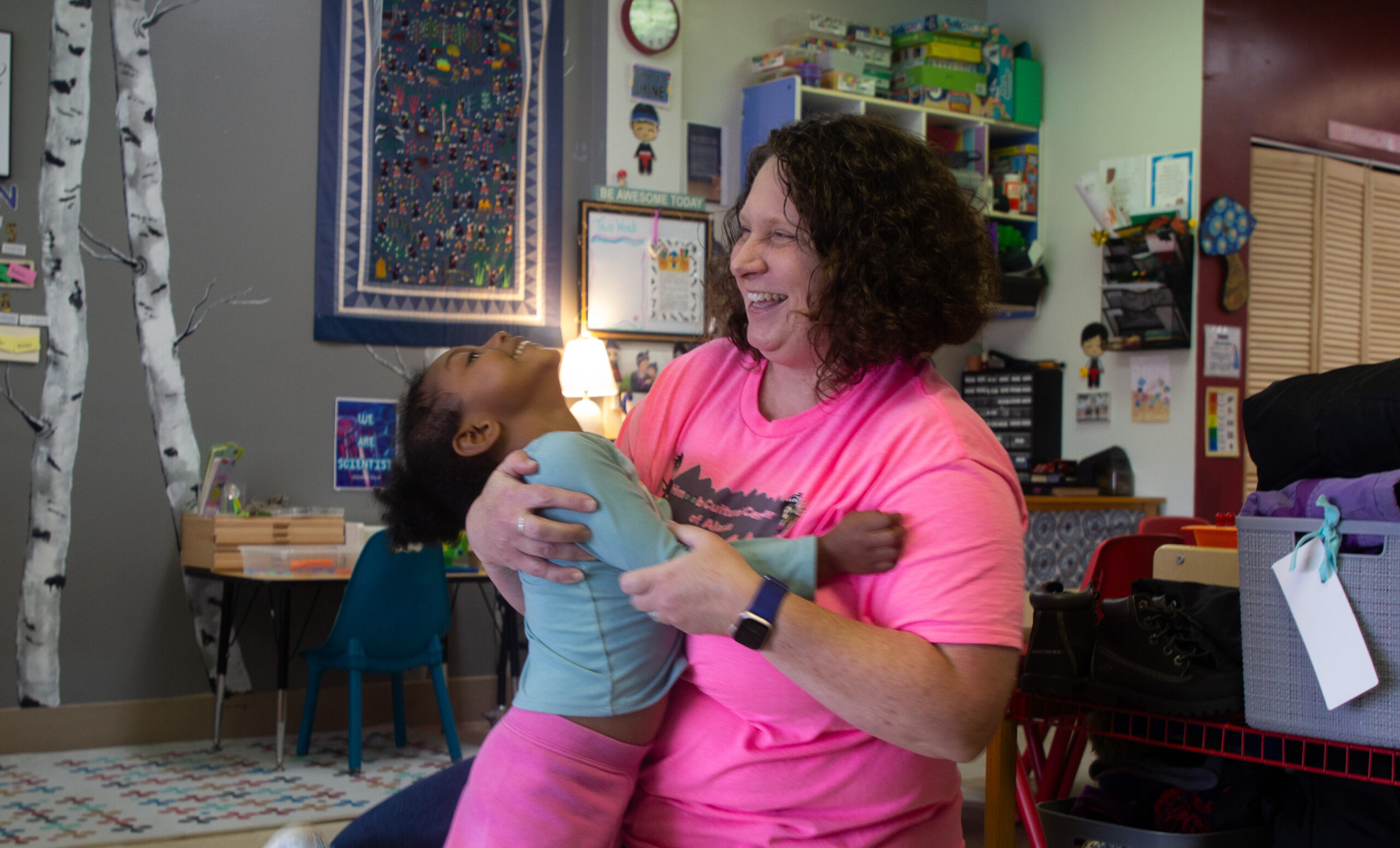 A smiling woman with brunette hair in a pink shirt holds a smiling child in a hug while kneeling on the ground.