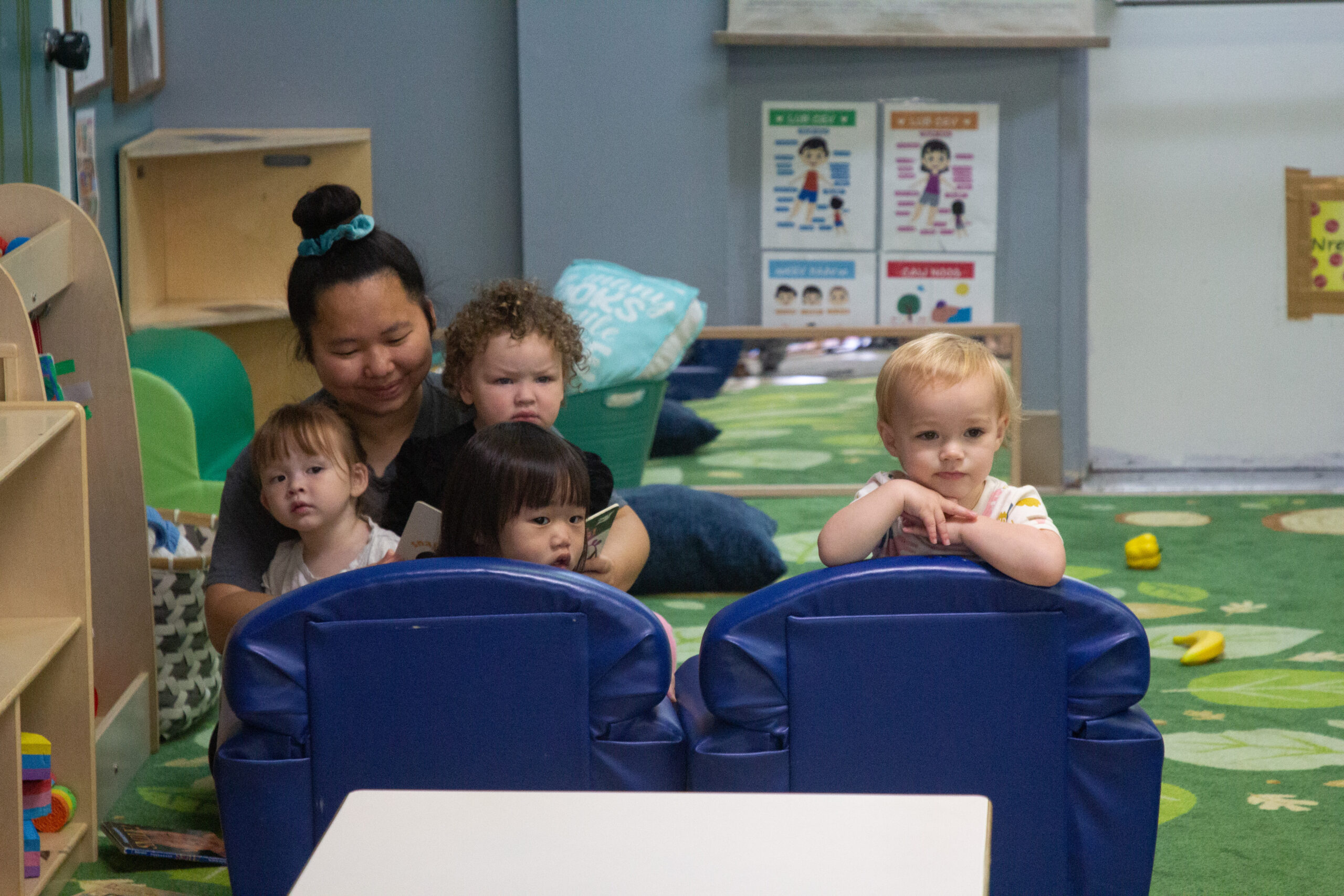 Four kids, two sitting in a chair look at something across the room as a lady in a grey shirt reads a story to them.