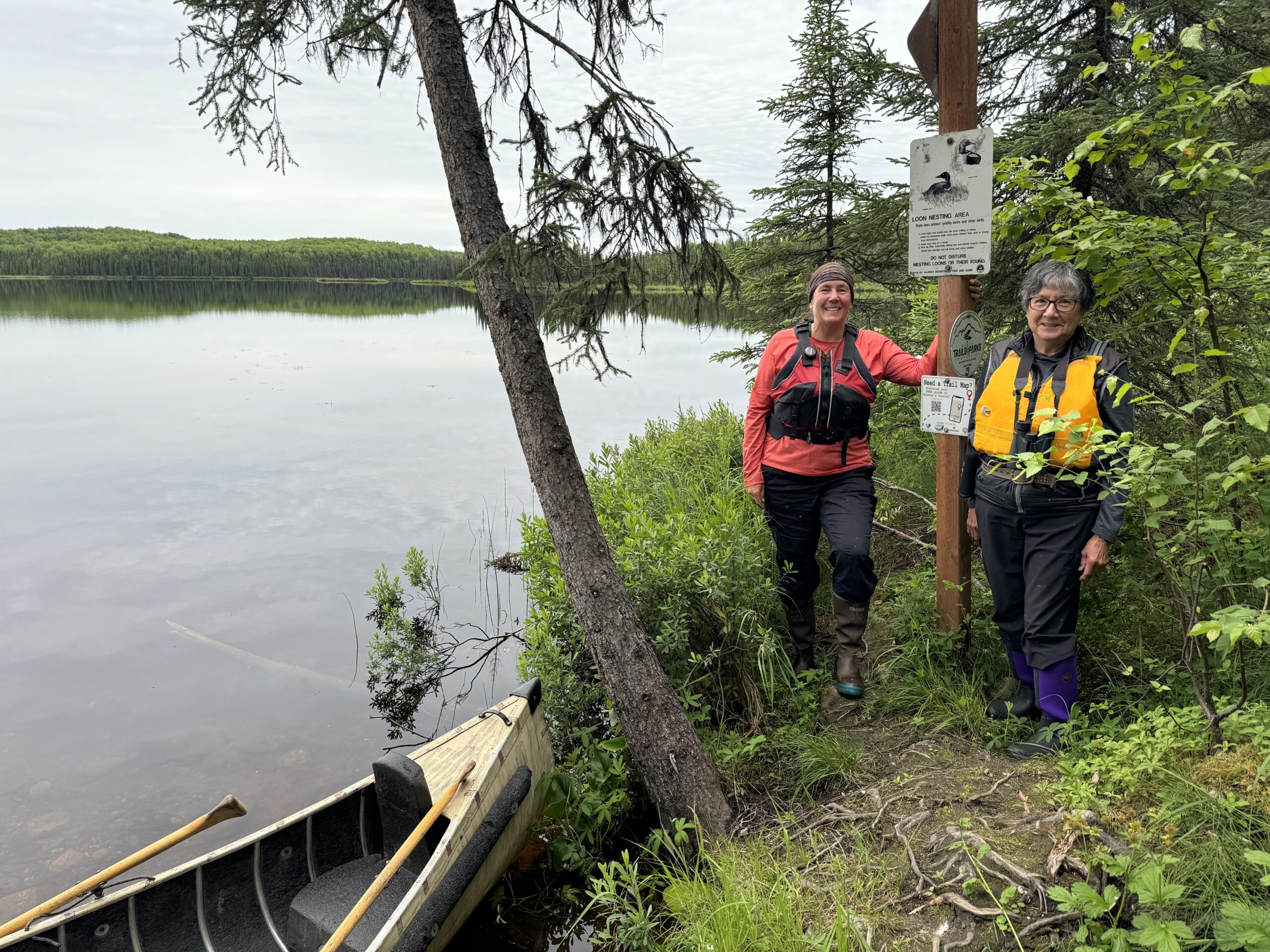 Two women successful life jackets guidelines adjacent to a canoe by a lake. 