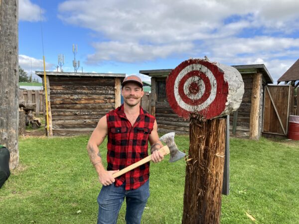 A lumberjack stands adjacent to a throwing axe target while holding a double sided axe. 
