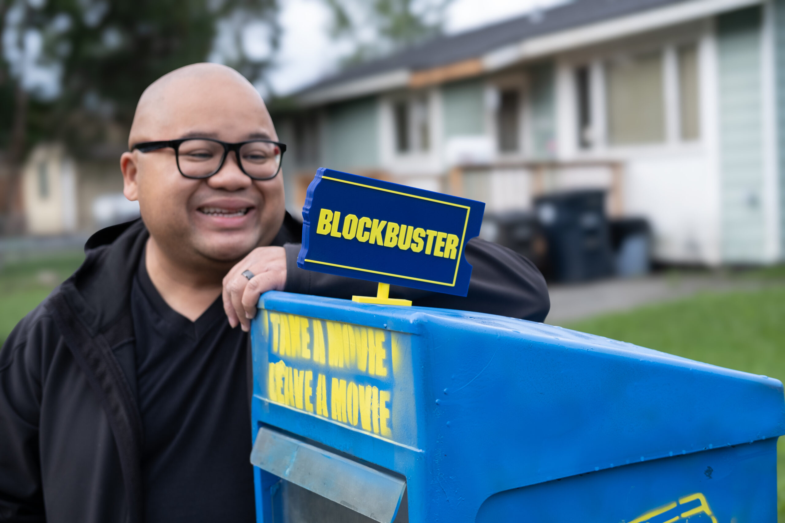 a small sign that says "blockbuster" is perched atop a blue box. a man with glasses is smiling behind it with a house in the far background