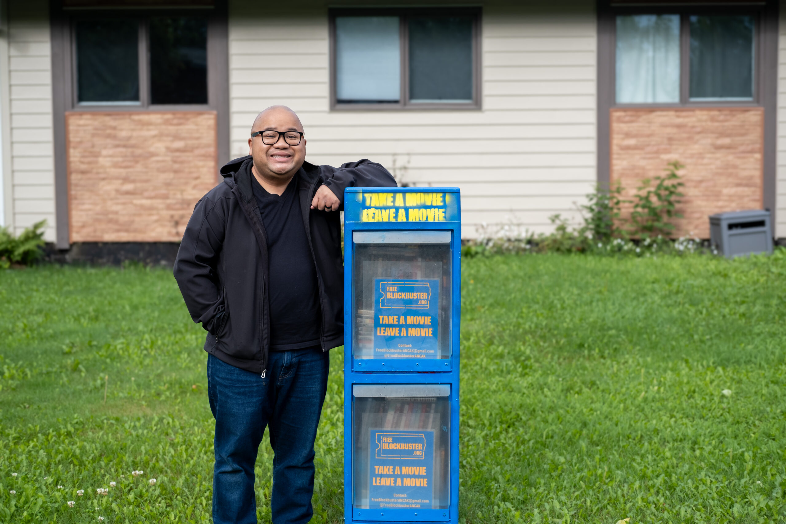 a man put his elbow on top of a blue newspaper box in the front yard of a house. he is smilling