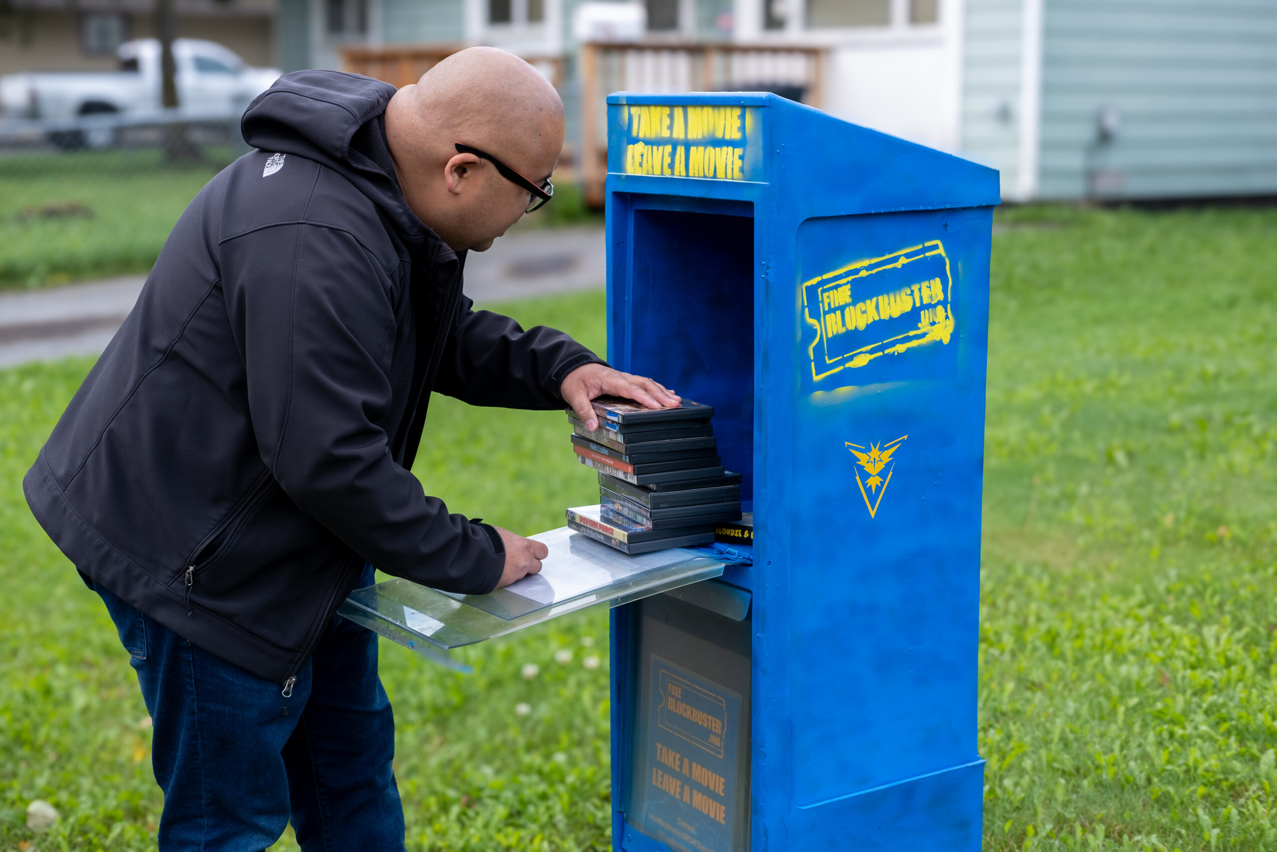 a man in a black jacket pulls DVD cases out of a blue newspaper box with the words "free block buster" and "take a movie leave a movie" spraypainted on the side