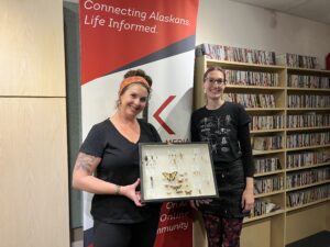 Two women hold a display of insect specimens. 