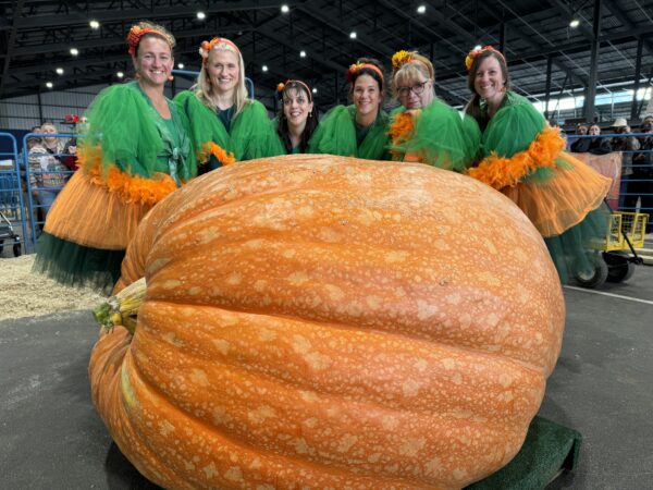 Women in costume stand behind a giant pumpkin. 