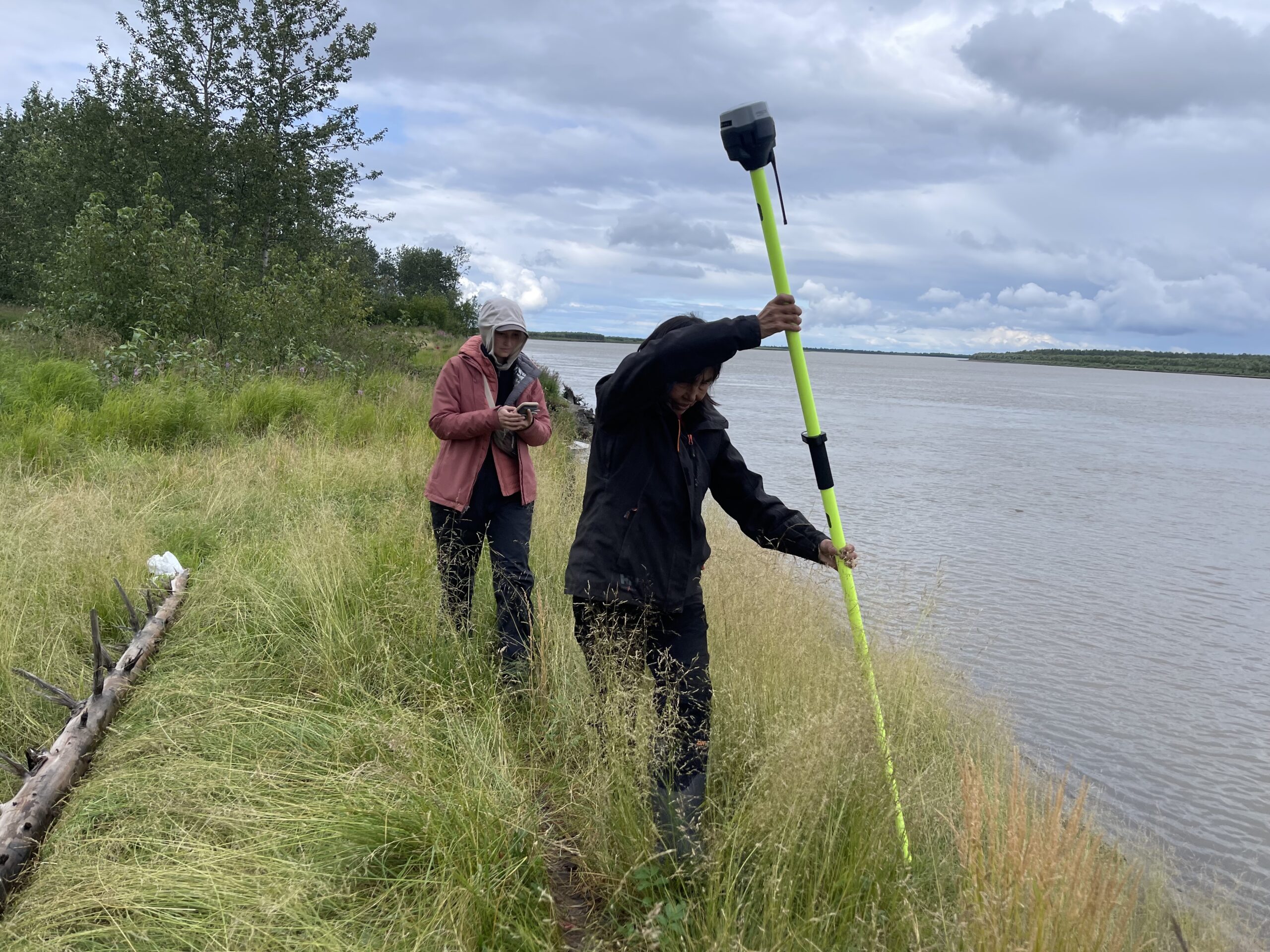 two women stand on the edge of a river bank. the woman in front is holding a very tall green pole with a grey box on top of it