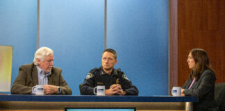 Two men, one in a suit and another in a police uniform, sit at a desk with a woman wearing a blazer.