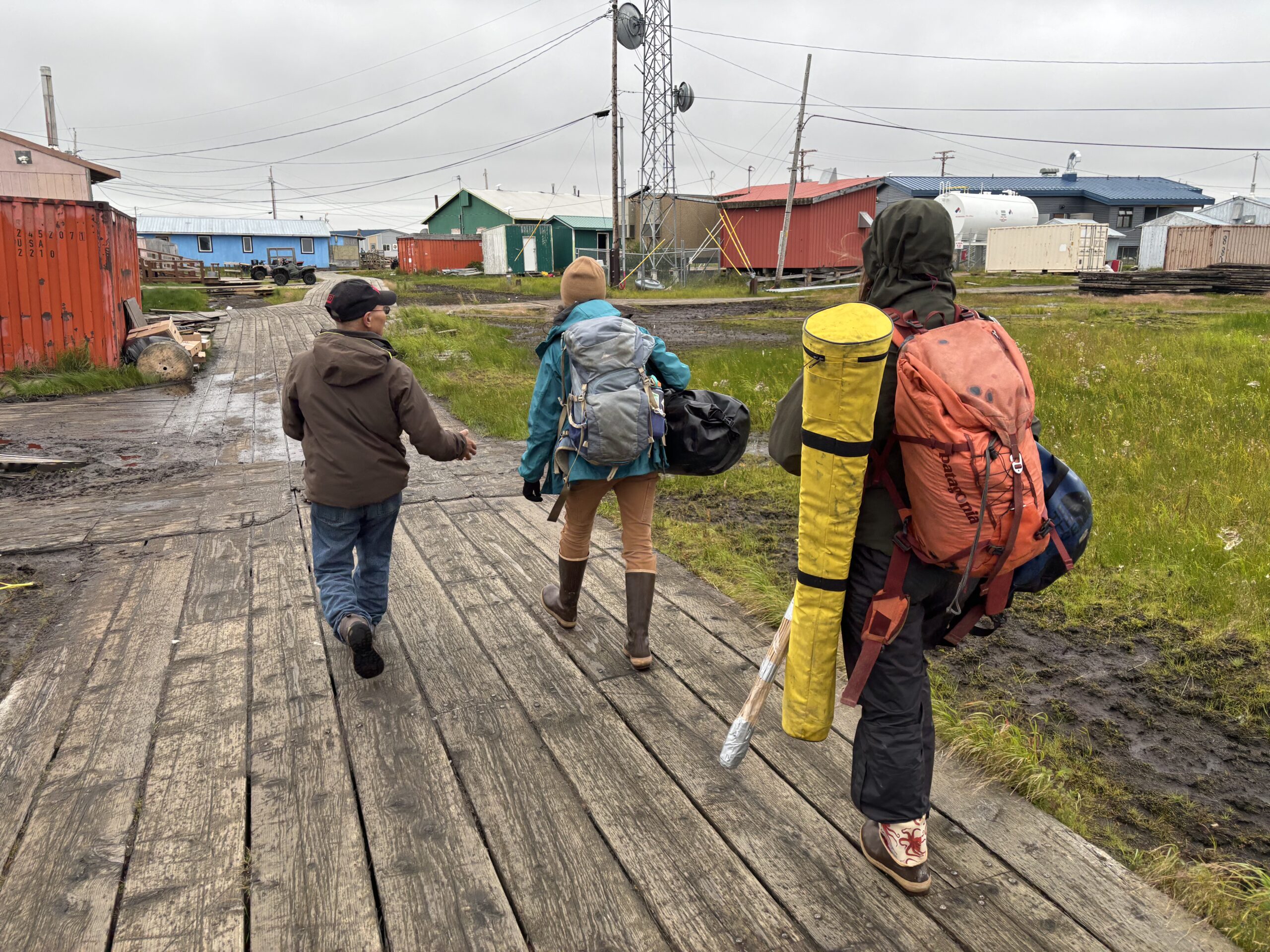 three people, 2 of whom are carrying large backpacks, locomotion on a wide boardwalk. location are colorful buildings successful beforehand of them.