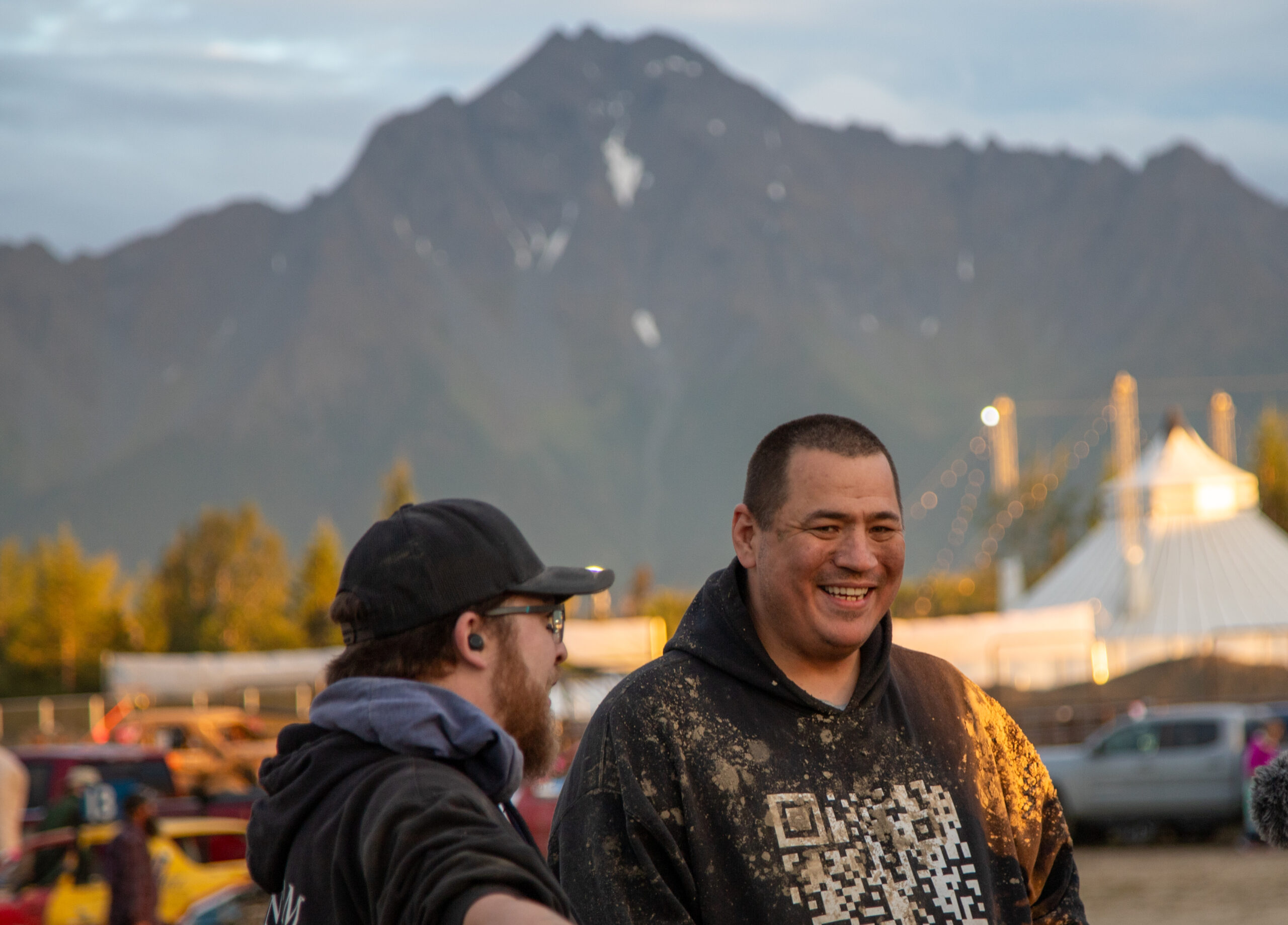 two men, both wearing black hoodies stand smiling with cars, a large tent, and a mountain in the background.