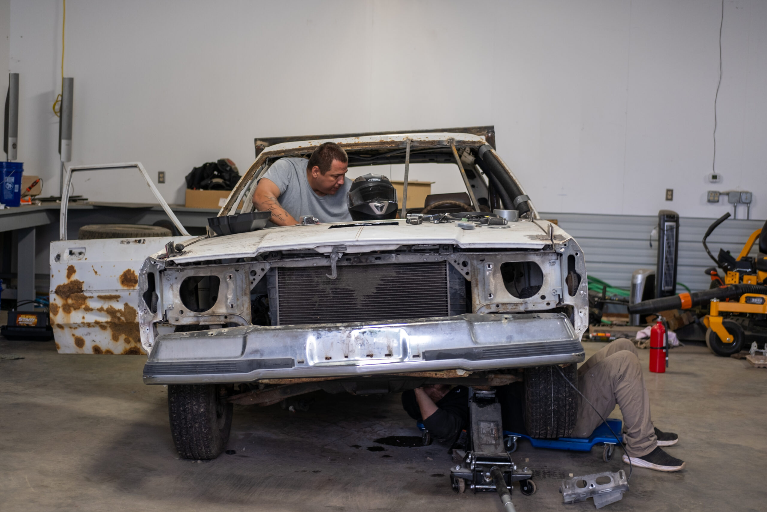 a man pops his head out of the windshield of a white car, looking focused. under the car you can see a different man's knees as he works on the underside of the car