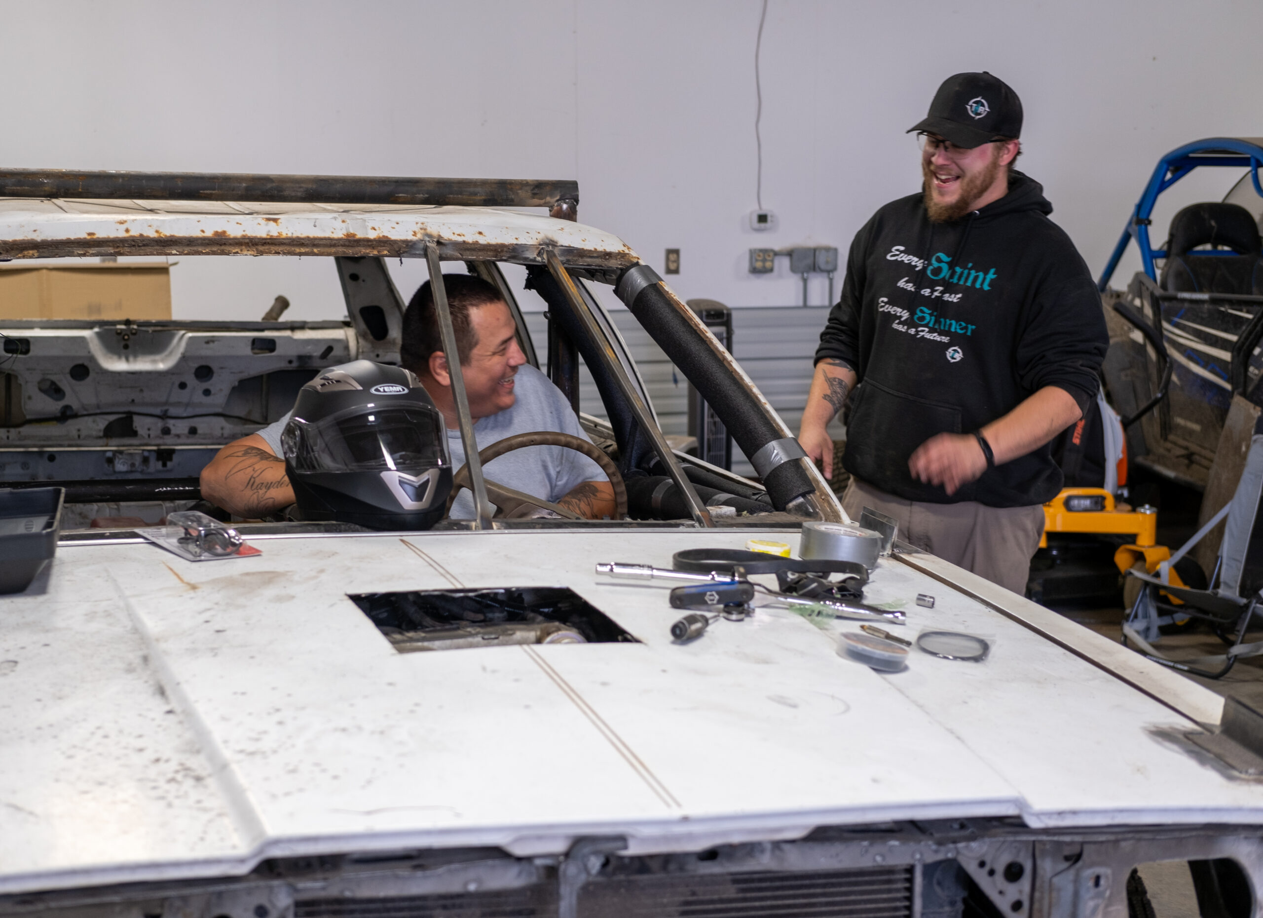 two men laugh together while working on a car in a garage