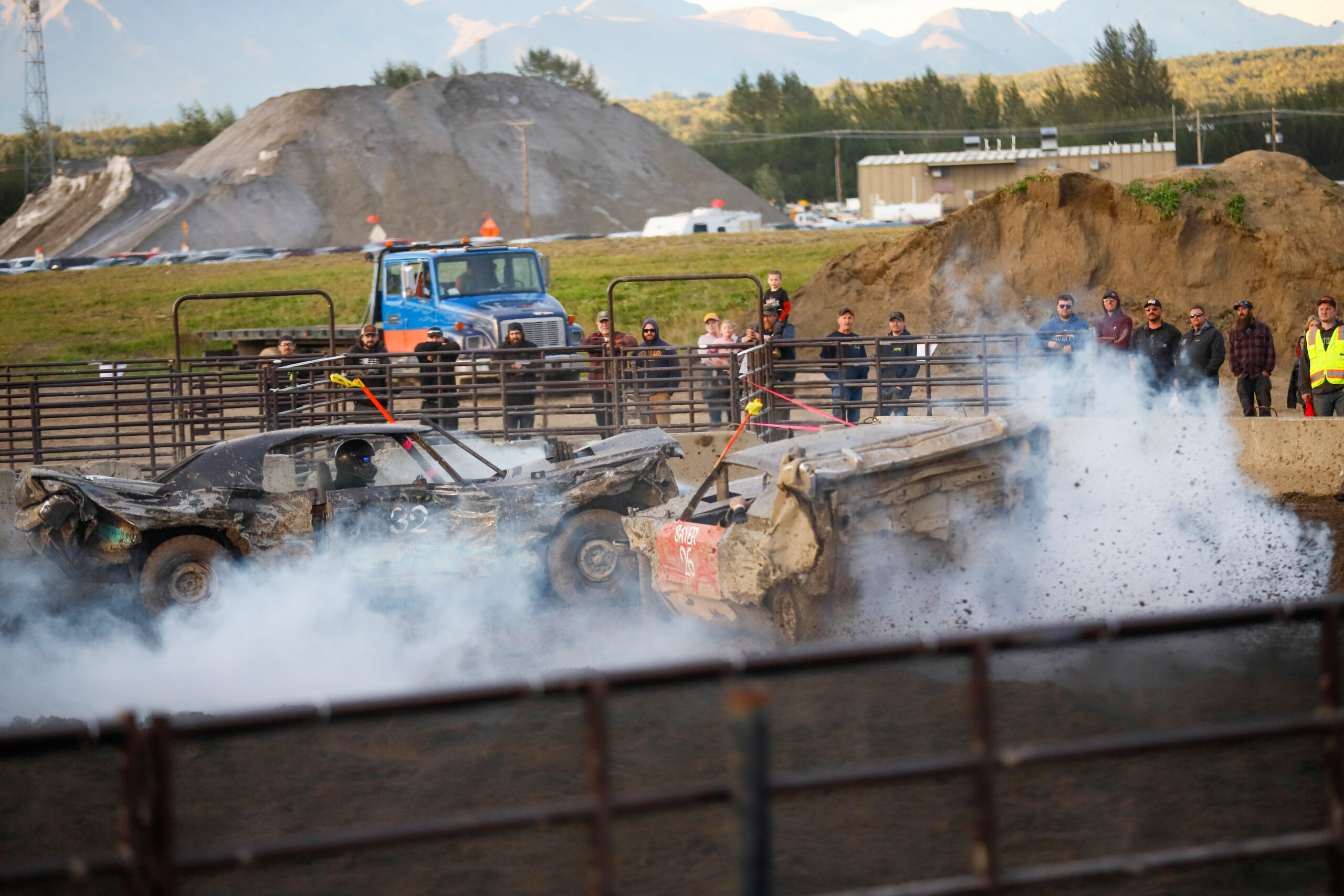 two cars clang into each other, some are very beaten up. a unreality of fume engulfs some of them and spectators watch from down a fence