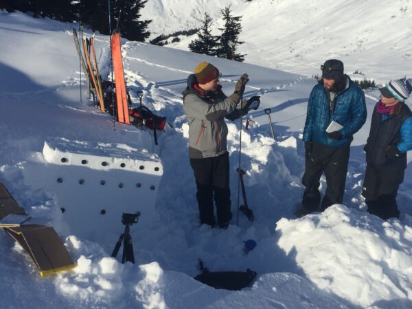 People stand on a snowy hill being told about the layers of snow to gauge avalanche risk.  