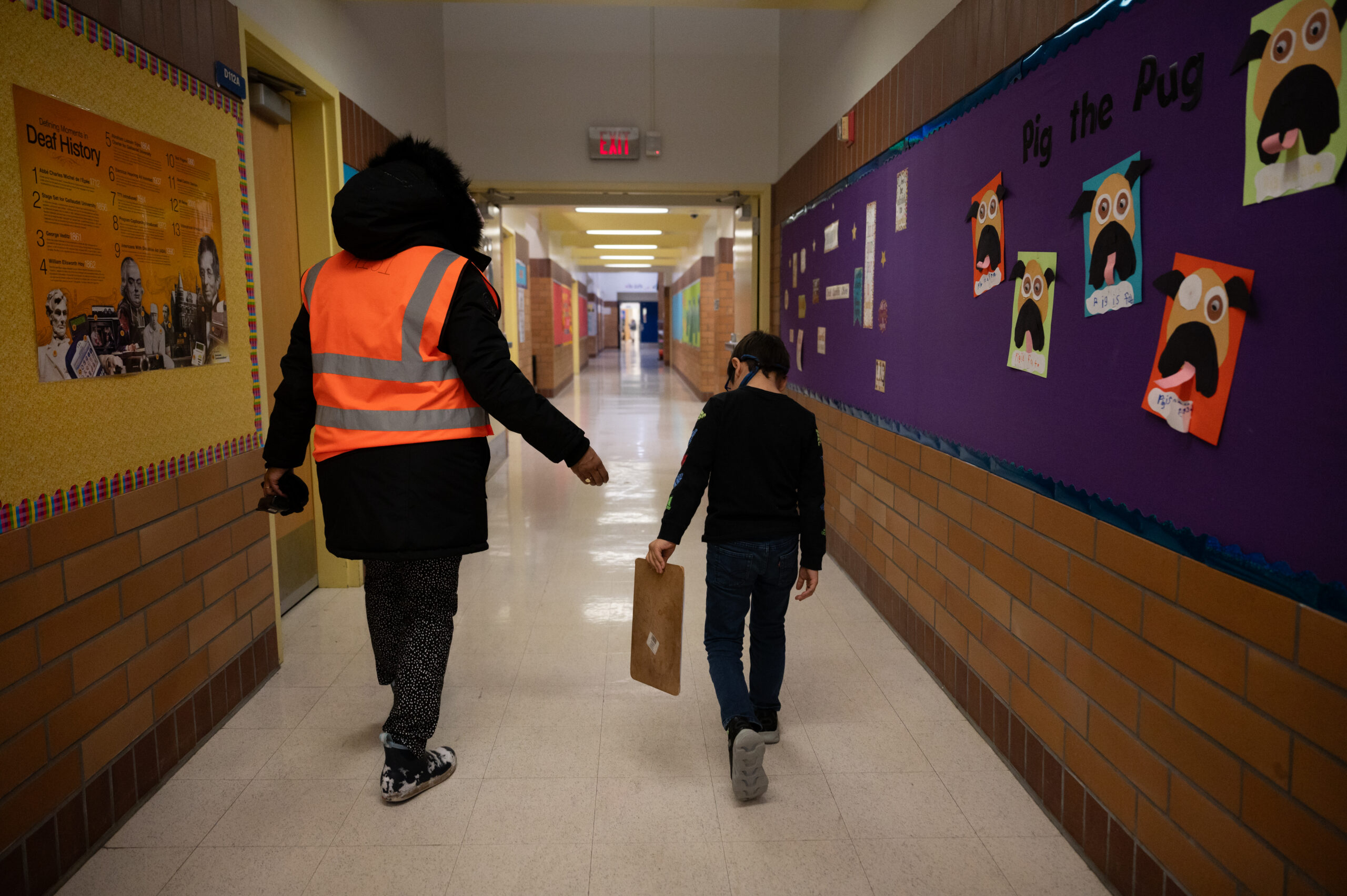 A woman in an orange safety vest helps a student down the hallway.