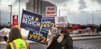 A man in a jacket looks at the camera holding a sign against ranked choice voting. While others in support of ballot initiative two and Nick Begich's campaign stand near the road with signs.