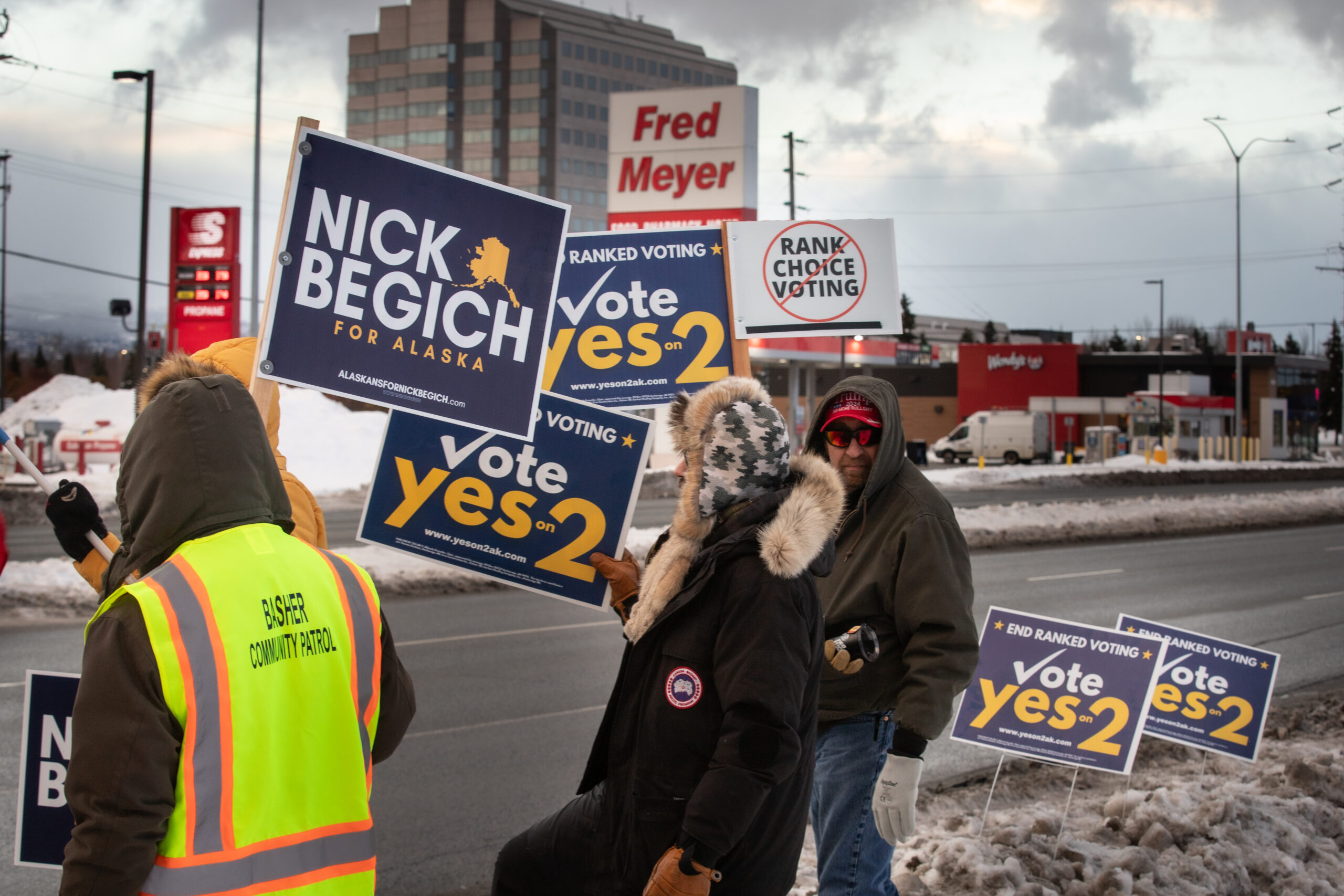 A man in a jacket looks at the camera holding a sign against ranked choice voting. While others in support of ballot initiative two and Nick Begich's campaign stand near the road with signs.