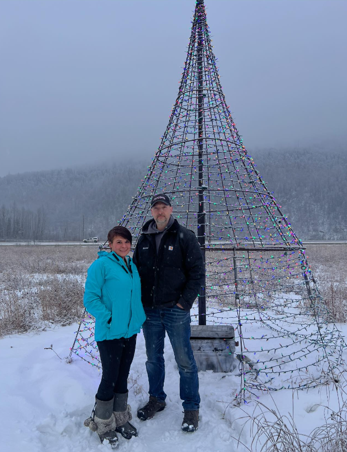 A man and woman stand in front of a metal Christmas tree in the snow.